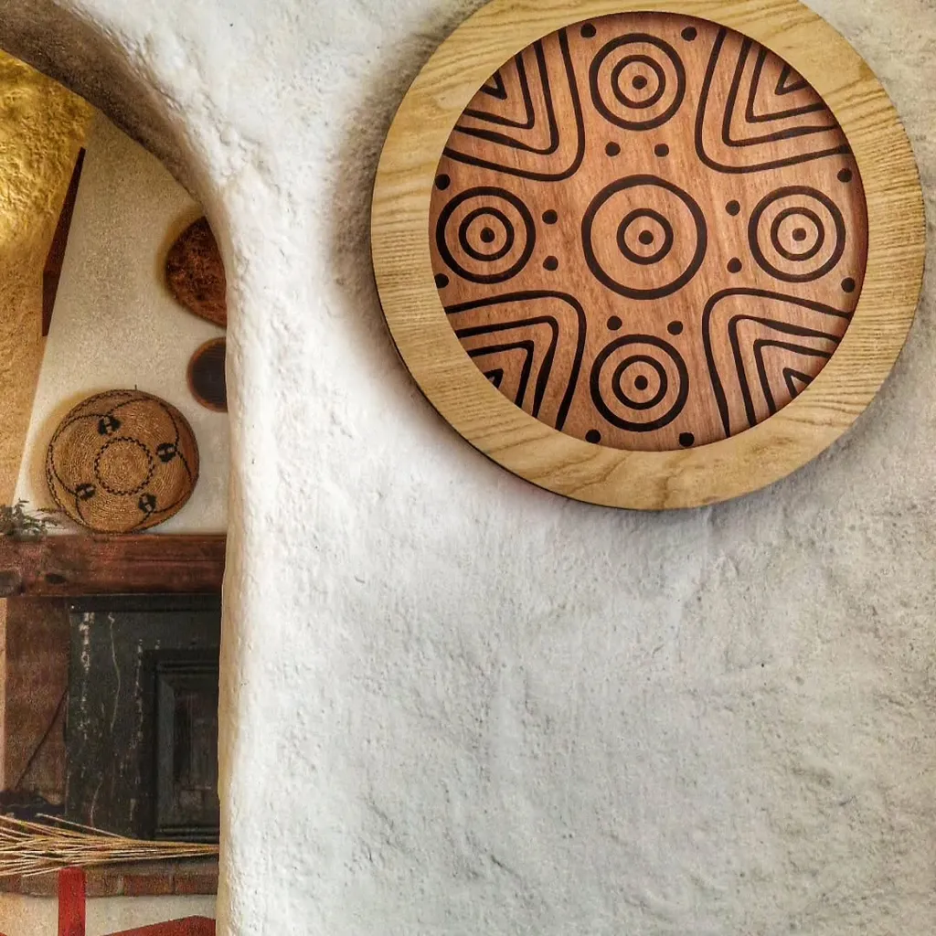 A close-up shot of a round wooden decorative piece with engraved abstract patterns, mounted on a textured white wall. A rustic fireplace with decorative baskets is partially visible on the left side of the frame.