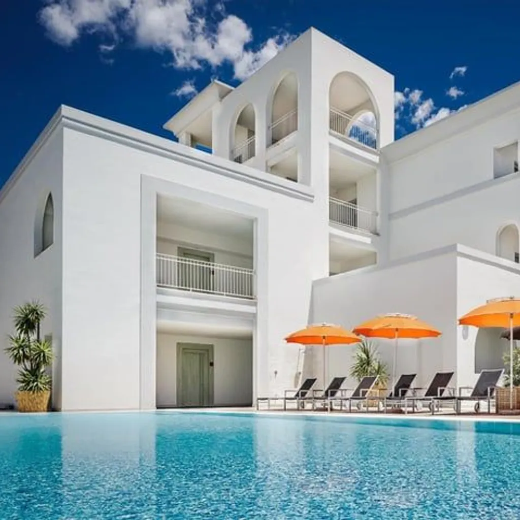 The image displays a well-composed scene of a modern white architectural building next to a vibrant blue swimming pool, adorned with orange sun umbrellas and lounge chairs under a clear blue sky. The lighting is bright and natural, highlighting the clean lines of the building and the sparkling water.