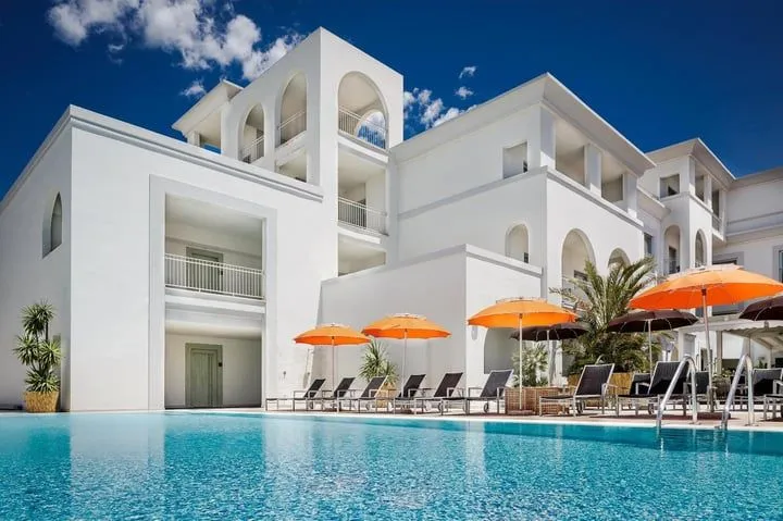 The image displays a well-composed scene of a modern white architectural building next to a vibrant blue swimming pool, adorned with orange sun umbrellas and lounge chairs under a clear blue sky. The lighting is bright and natural, highlighting the clean lines of the building and the sparkling water.