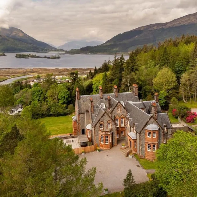 An aerial view of a grand stone building surrounded by lush green trees, with a large loch or lake and mountains in the background under a cloudy sky. The building appears to be a historic mansion or hotel, blending well with the natural scenery.