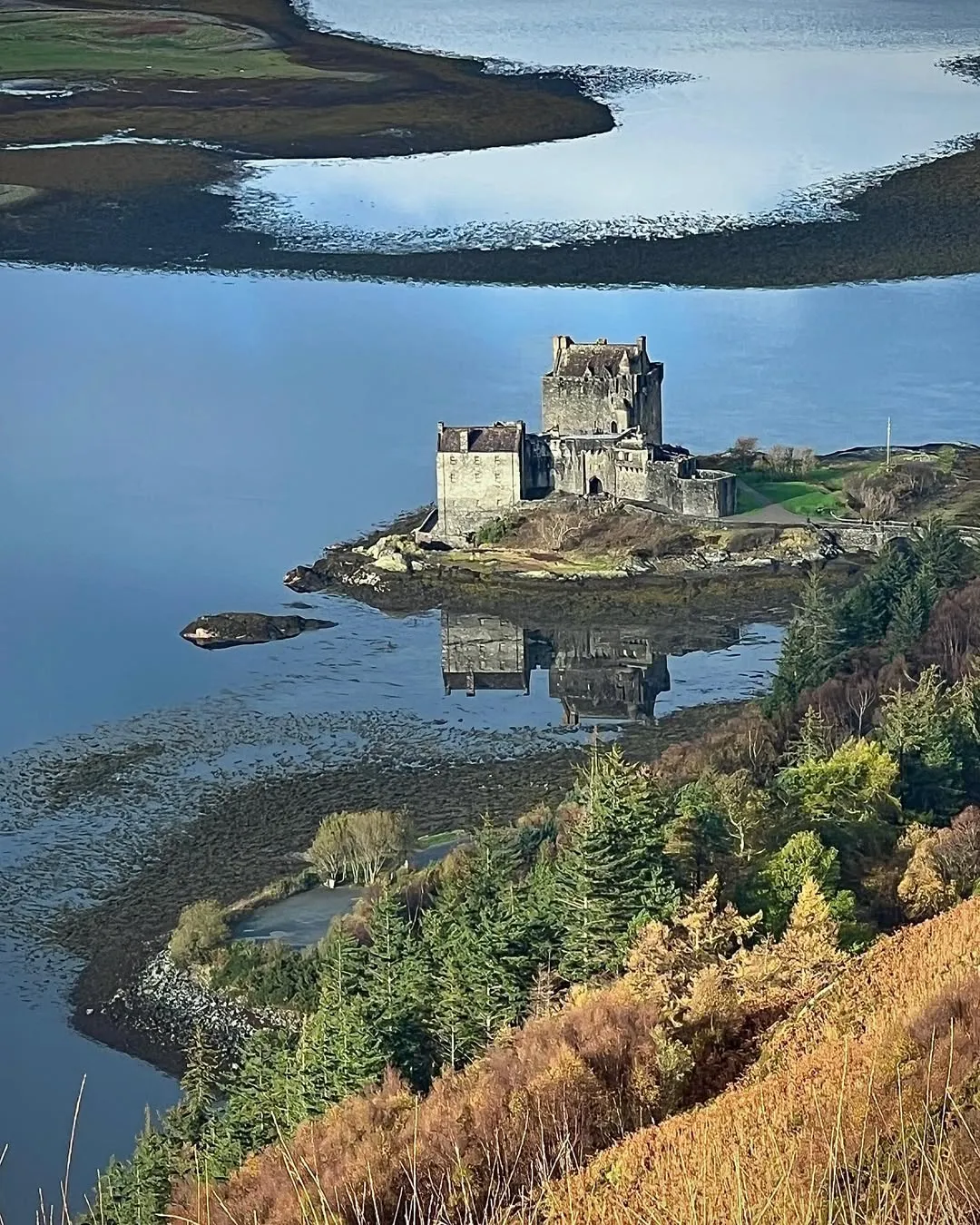 The image captures Eilean Donan Castle beautifully, showcasing its iconic position surrounded by water and autumnal foliage. The composition is strong, with the castle prominently featured and its reflection adding depth to the scene. The lighting is natural and even, highlighting details without harsh shadows. Colors appear accurate and realistic, contributing to the overall photo-realistic quality. The sharpness is good, with the main subject and foreground elements in clear focus.