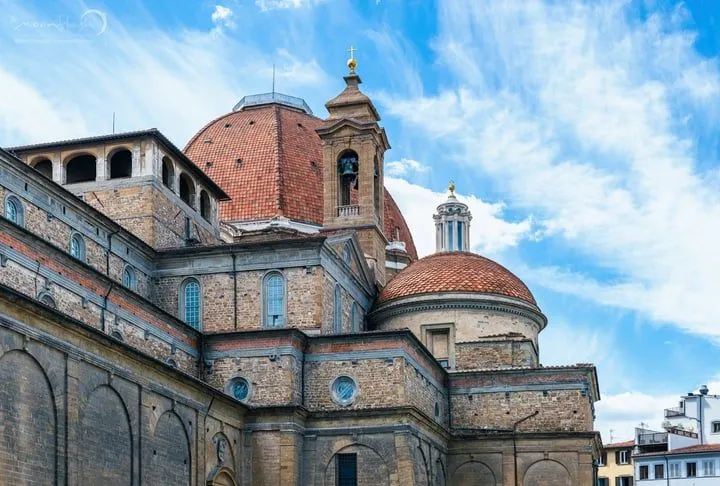 An editorial style image showcasing the architectural details of a historic building, likely a cathedral or basilica, with prominent domes and a bell tower against a blue sky with clouds.