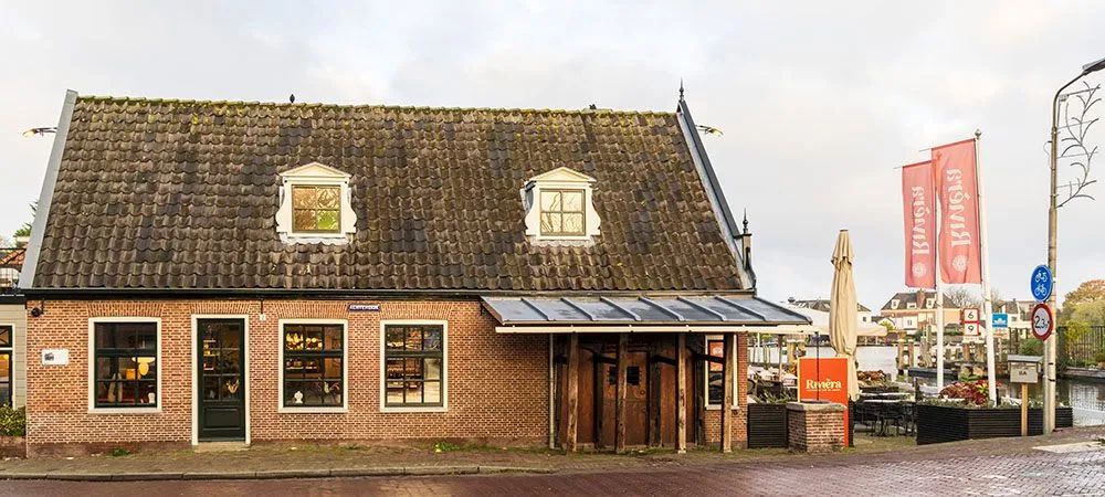 An exterior shot of an old brick building with a tiled roof, multiple windows, and a prominent wooden entrance. The image is well-composed and sharp, capturing good detail of the building's facade. The lighting is natural but appears somewhat flat due to overcast conditions, which slightly diminishes the overall vibrancy.