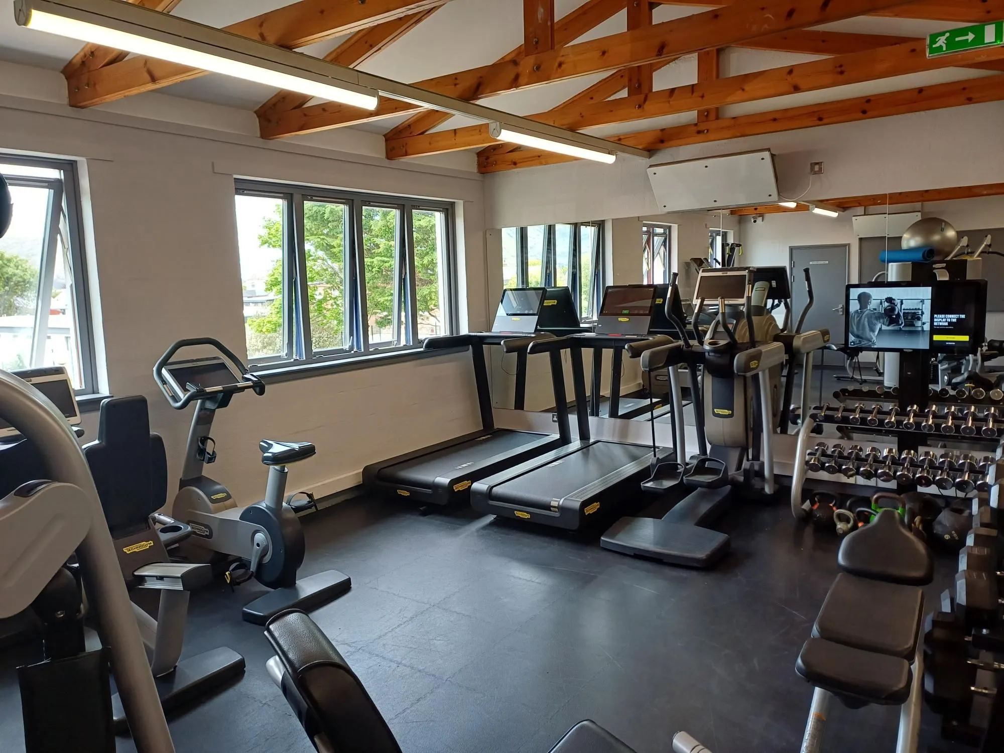 An interior shot of a bright and well-equipped gym featuring treadmills, an exercise bike, and weight racks. The space is characterized by large windows and exposed wooden ceiling beams.