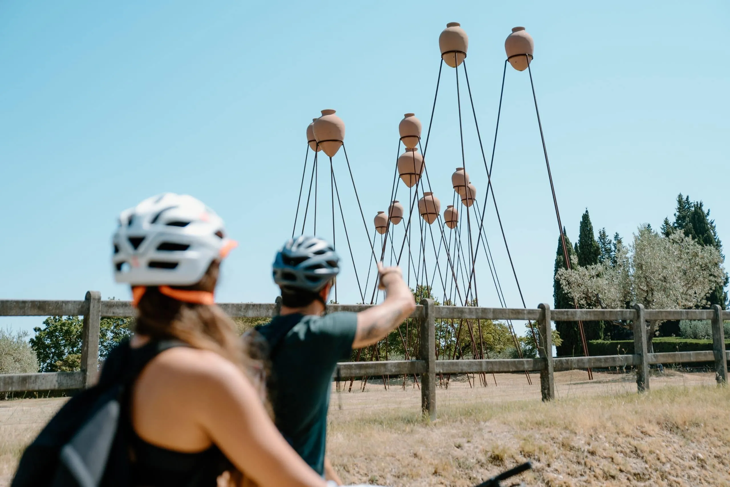 An outdoor, editorial-style image featuring two individuals on bicycles viewing a unique art installation of terracotta pots on tall stilts against a clear blue sky. The scene is set in a natural, open environment with a wooden fence and trees in the background.