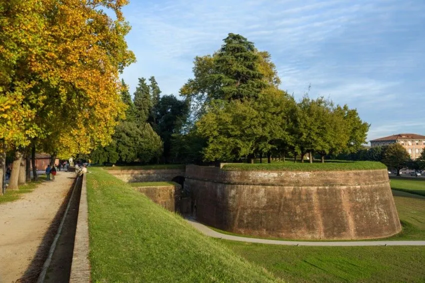 An editorial-style image featuring a curved ancient brick wall, likely part of a city fortification, surrounded by well-maintained green grass and various trees under a bright blue sky.