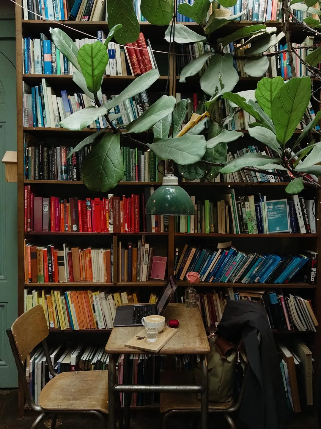 A cozy interior scene featuring a large wooden bookshelf filled with numerous books, accented by a lush green plant. A small wooden table with a laptop, a cup of coffee, and a vintage green lamp creates an inviting workspace.