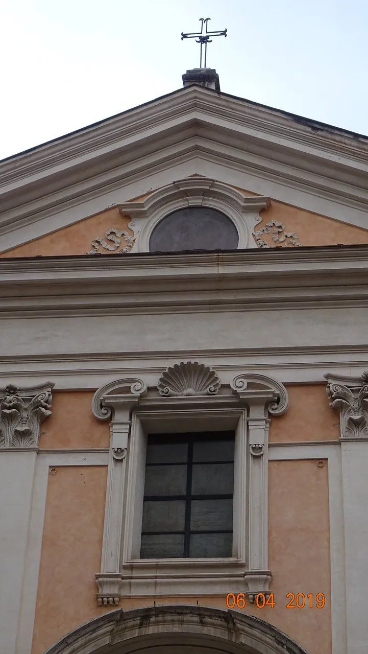 A detailed, editorial-style close-up of a building facade, showcasing classical architectural elements such as a pediment, an oculus window, and a lower window adorned with ornate shell and scroll motifs. The structure features light-colored stone or plaster with contrasting pale orange/peach sections and intricate carvings.