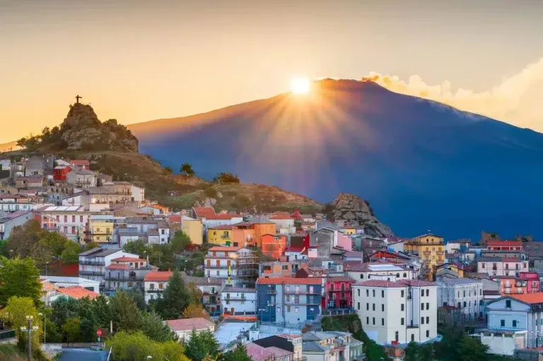 An aerial view of a colorful village nestled on a hillside, with a majestic mountain in the background bathed in the warm glow of a setting or rising sun, creating dramatic sun rays.