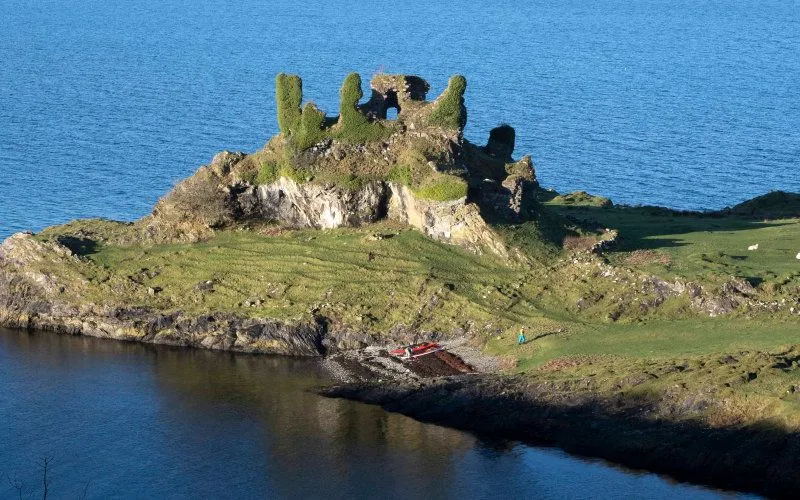 A wide aerial shot of a small, rocky island featuring ancient, overgrown castle ruins, surrounded by calm, deep blue water. The island has patches of green grass and a small, sheltered cove with some red debris and a tiny person visible on the shore.