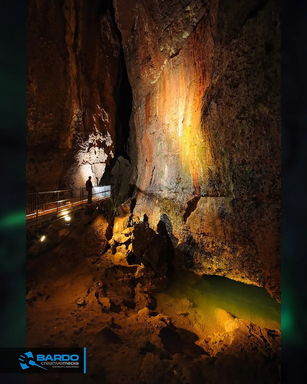 An image capturing the interior of a cave, featuring a person on a well-lit walkway, intricate rock formations, and a small pool of water. The scene is illuminated by artificial lighting, highlighting the natural textures and colors of the cave.