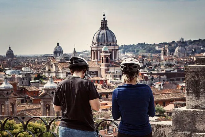 An editorial-style image depicting two individuals wearing bike helmets, seen from behind, looking out over a historic cityscape. A prominent dome structure dominates the background. The composition effectively frames the city view with the subjects in the foreground.