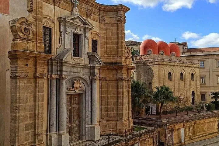 A vibrant architectural scene featuring ornate historic stone buildings under a bright blue sky, with distinctive red domes in the background.