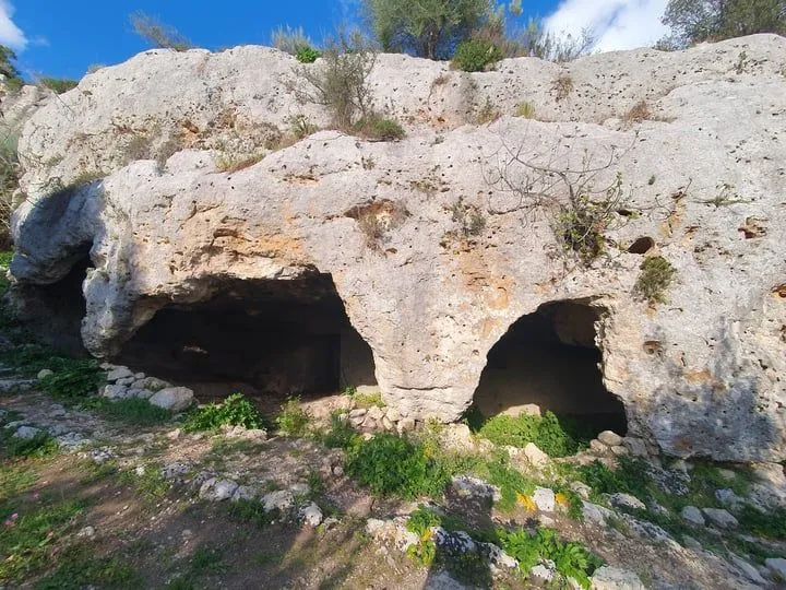An editorial style image showcasing two dark cave entrances within a natural rocky cliff formation, surrounded by sparse vegetation under a blue sky.