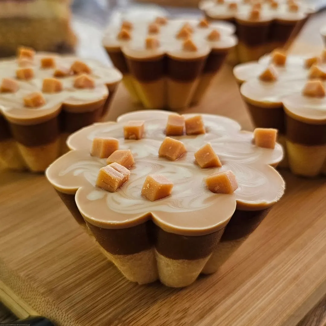 The image showcases several individual caramel desserts on a wooden surface. The foreground dessert is in sharp focus, highlighting the distinct layers and the small caramel cubes scattered on top of a swirled light-colored topping. The other desserts are softly blurred in the background, creating a pleasant depth of field. The lighting is even and soft, making the textures and colors of the desserts appealing and realistic. The composition effectively draws attention to the main subject, making it look delicious and professionally presented.