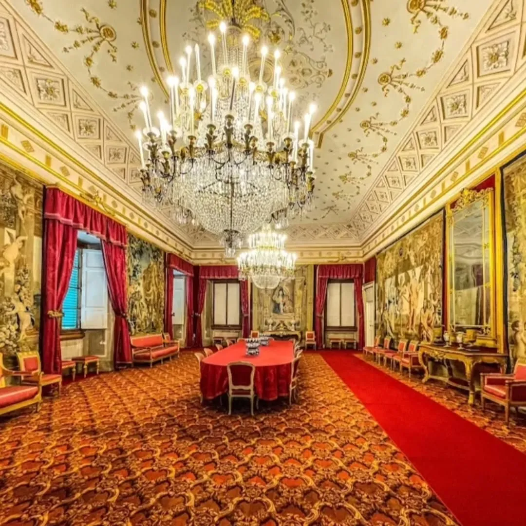 An opulent and grand hall or dining room, likely within a palace or historical building, featuring a long red-draped table, intricate chandeliers, vibrant red carpet, and richly decorated walls with tapestries. The ceiling is ornate with gold accents.