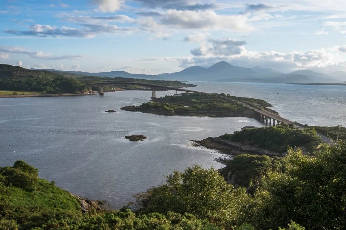 This image showcases a well-composed and realistic landscape featuring a bridge over water, surrounded by hills and distant mountains under a partly cloudy sky. The lighting is natural and effective, contributing to the photo's overall clarity and depth. Key elements, including the bridge and foreground, are sharp and in focus, with accurate colors. The primary strength lies in its expansive and balanced composition, offering a highly photo-realistic view. Minor weaknesses include slight atmospheric softening of the most distant mountains, which is typical for such wide views, and some darker areas in the foreground foliage.