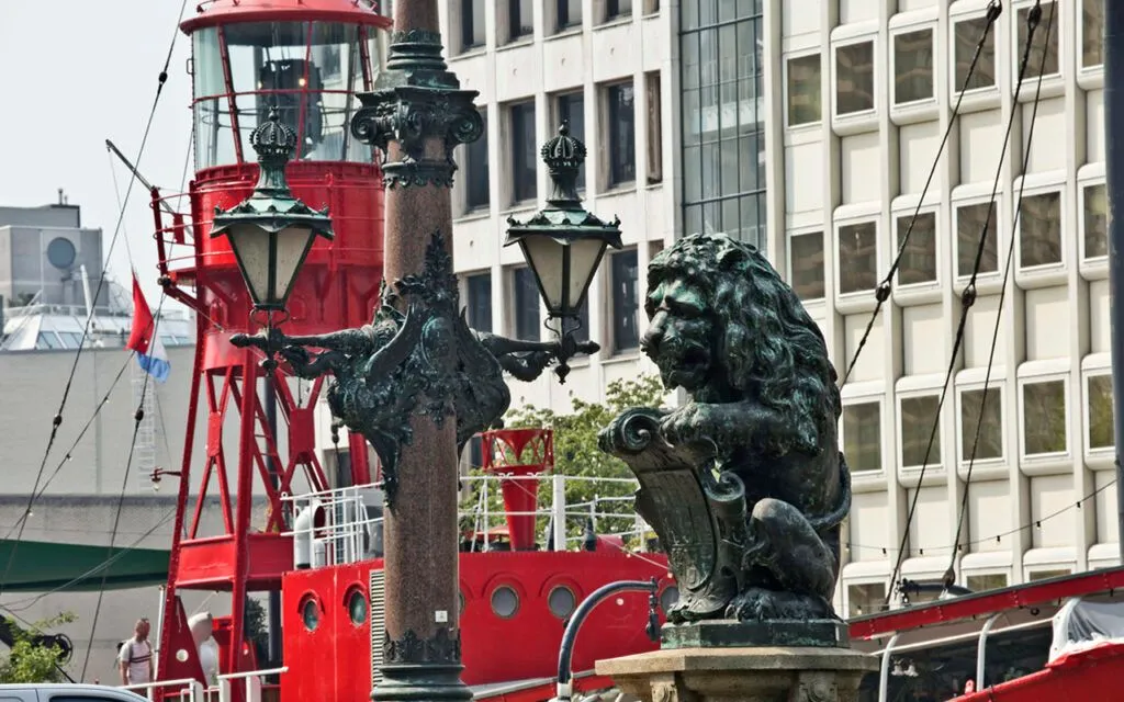 An image featuring a bronze lion statue and an ornate lamppost in the foreground, set against a backdrop of a red boat structure and a modern building. The composition is strong, balancing detailed foreground elements with interesting background textures and colors. The lighting is natural and even, highlighting the intricate details of the statue and lamppost. The image is sharp and photo-realistic, with accurate colors that enhance the scene's authenticity.