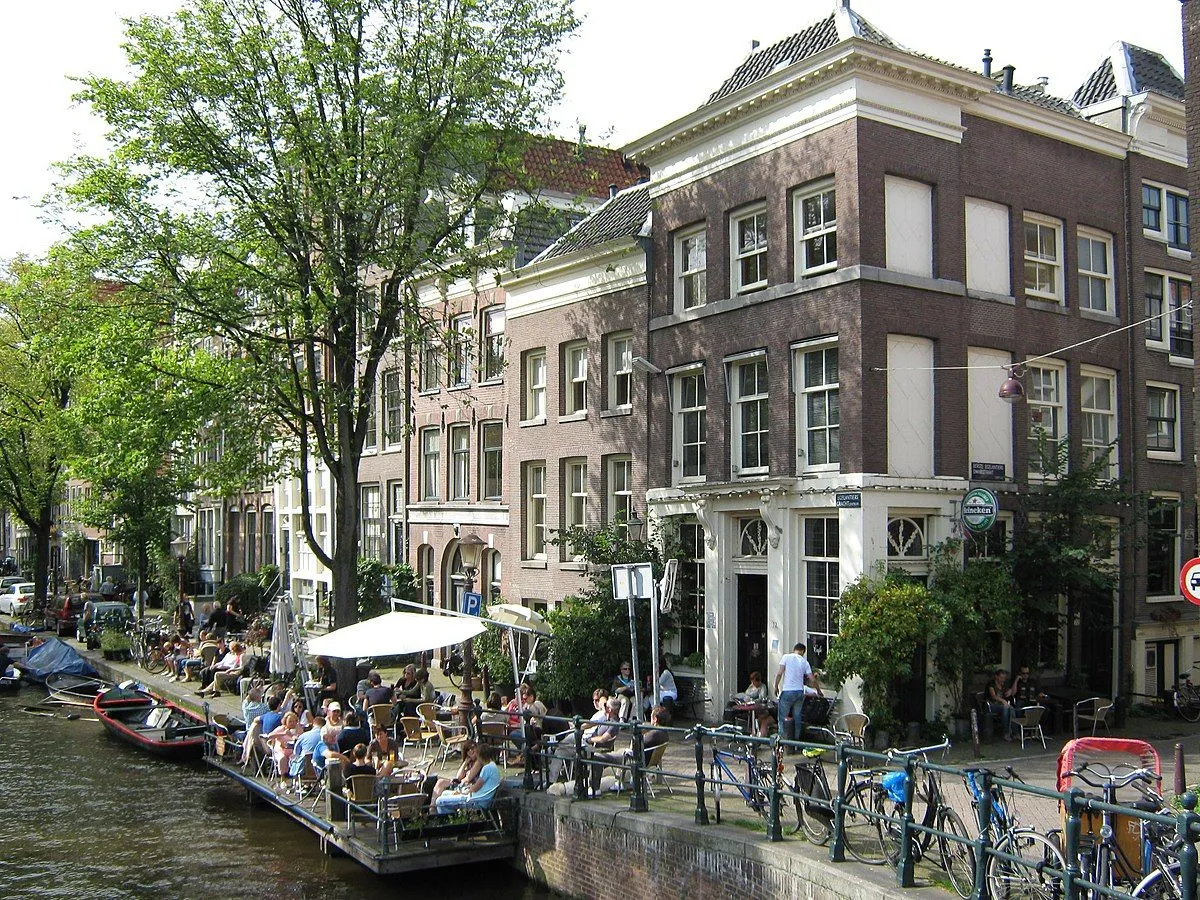 An outdoor cafe bustling with people along a canal, framed by traditional buildings and lush trees.