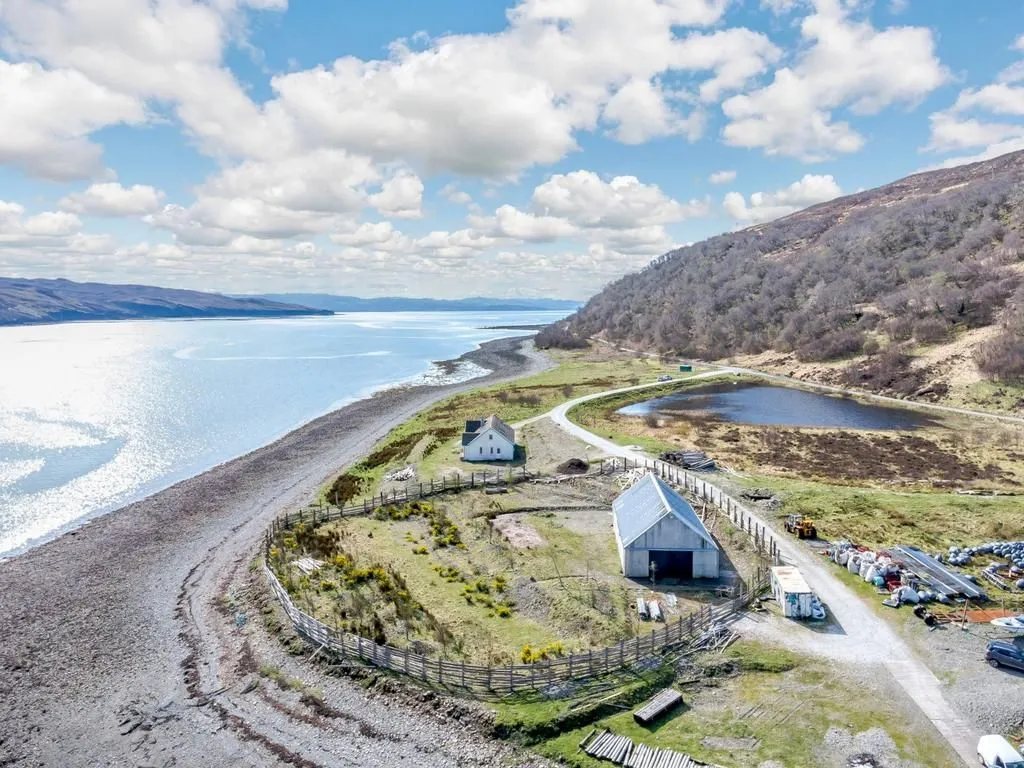 An aerial view captures a remote coastal landscape, featuring a large body of water, a winding road, and two isolated buildings set against a backdrop of hills and a cloudy sky.