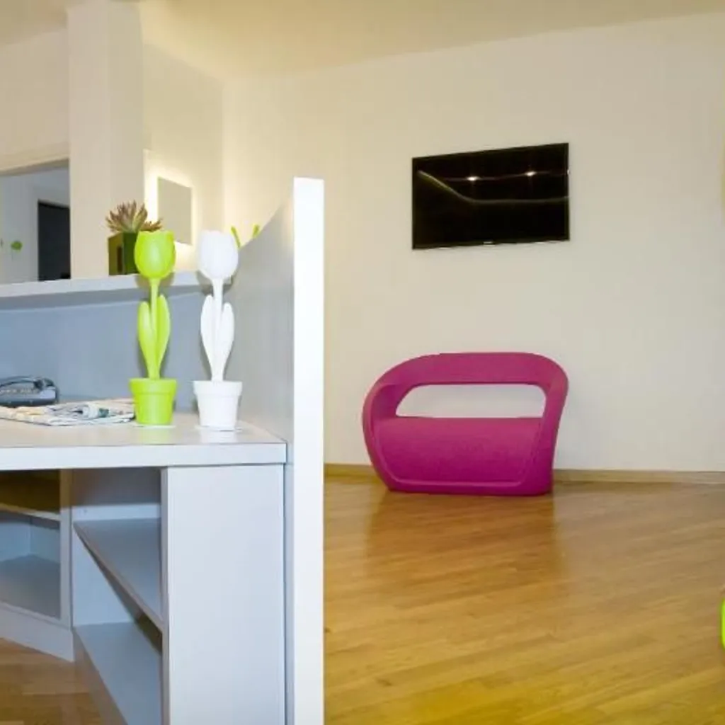 A well-lit interior shot featuring a desk with decorative tulip figures in the foreground, and a vibrant pink chair with a mounted television on the wall in the background. The room has light wooden flooring.