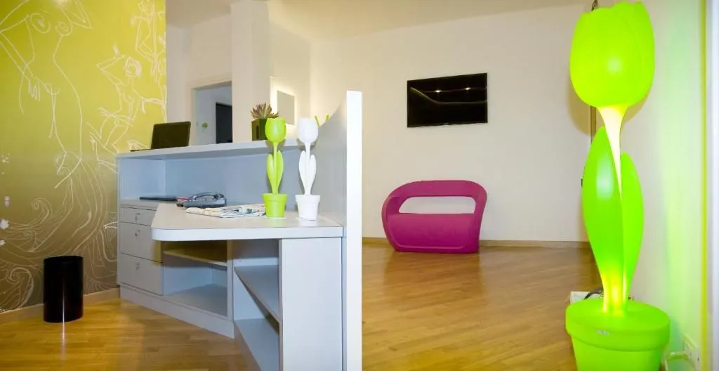 A well-lit interior shot featuring a desk with decorative tulip figures in the foreground, and a vibrant pink chair with a mounted television on the wall in the background. The room has light wooden flooring.
