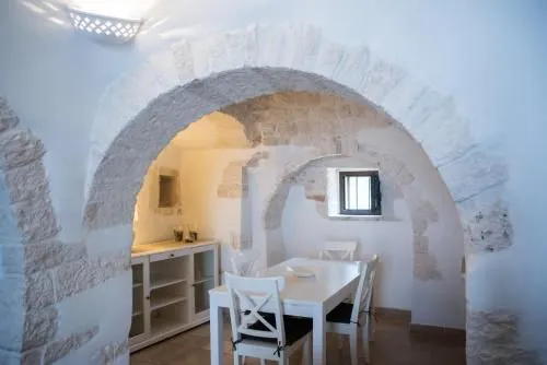 An interior shot showcasing a dining area and an adjacent sideboard, framed by multiple ancient stone arches. The architecture features exposed stone work contrasted with smooth white walls and white wooden furniture, typical of traditional Mediterranean or historic European dwellings.