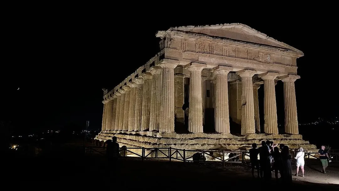 An ancient temple is captured at night, prominently illuminated against a dark sky. The image emphasizes the architectural details of the temple, with a few silhouetted individuals in the foreground providing a sense of scale.