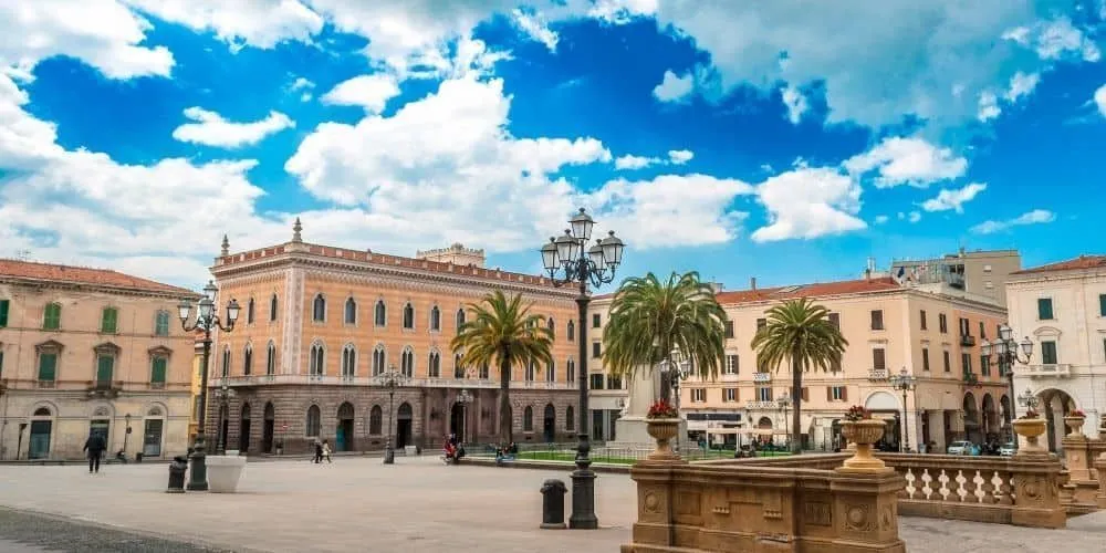An outdoor shot of a historic city square featuring a large, ornate building with classical architecture, palm trees, a prominent ornate lamppost, and a clear blue sky dotted with white clouds.