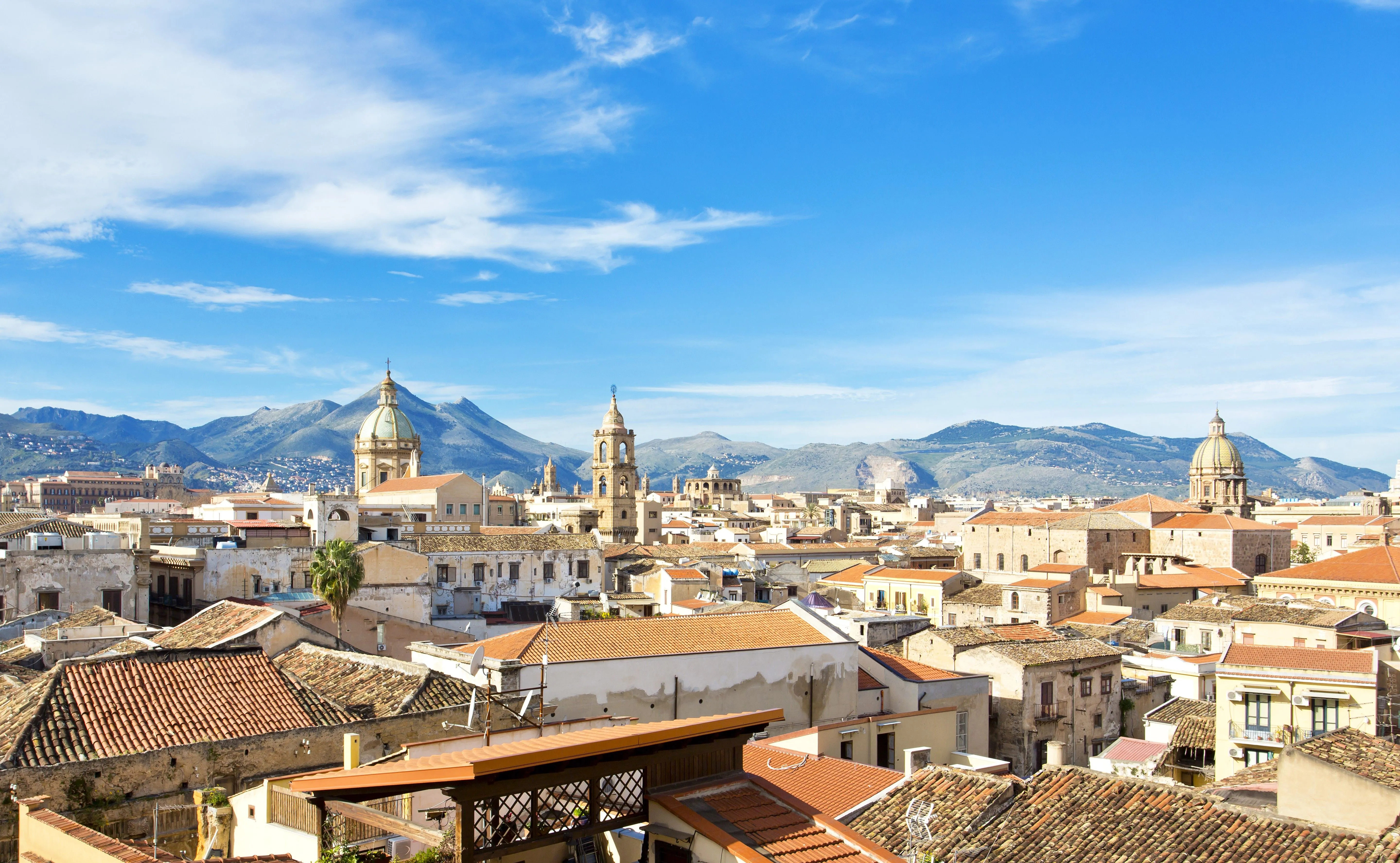 The image is well-composed with a clear hierarchy of elements, showcasing the city's architecture against a beautiful blue sky and distant mountains. Lighting is excellent, providing good visibility and detail without harsh shadows. The image is sharp throughout, particularly in the foreground and mid-ground structures, and colors appear natural and accurate. It exhibits strong photo-realism with all key elements in clear focus.
