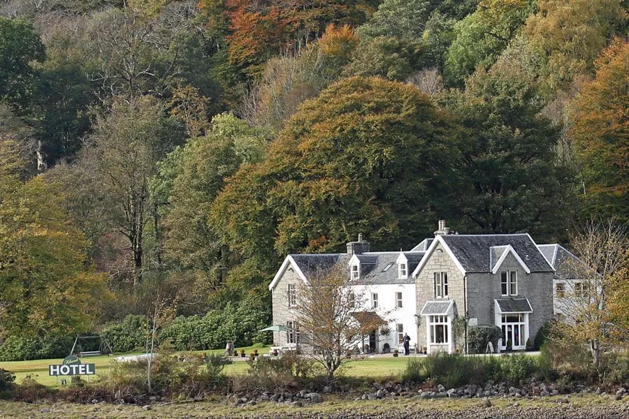A tranquil outdoor scene featuring a large, multi-story stone and white building, resembling a house or hotel, nestled by a rocky bank or shoreline. Behind the building rises a dense hillside forest displaying a mix of green and autumnal foliage, suggesting a fall season.