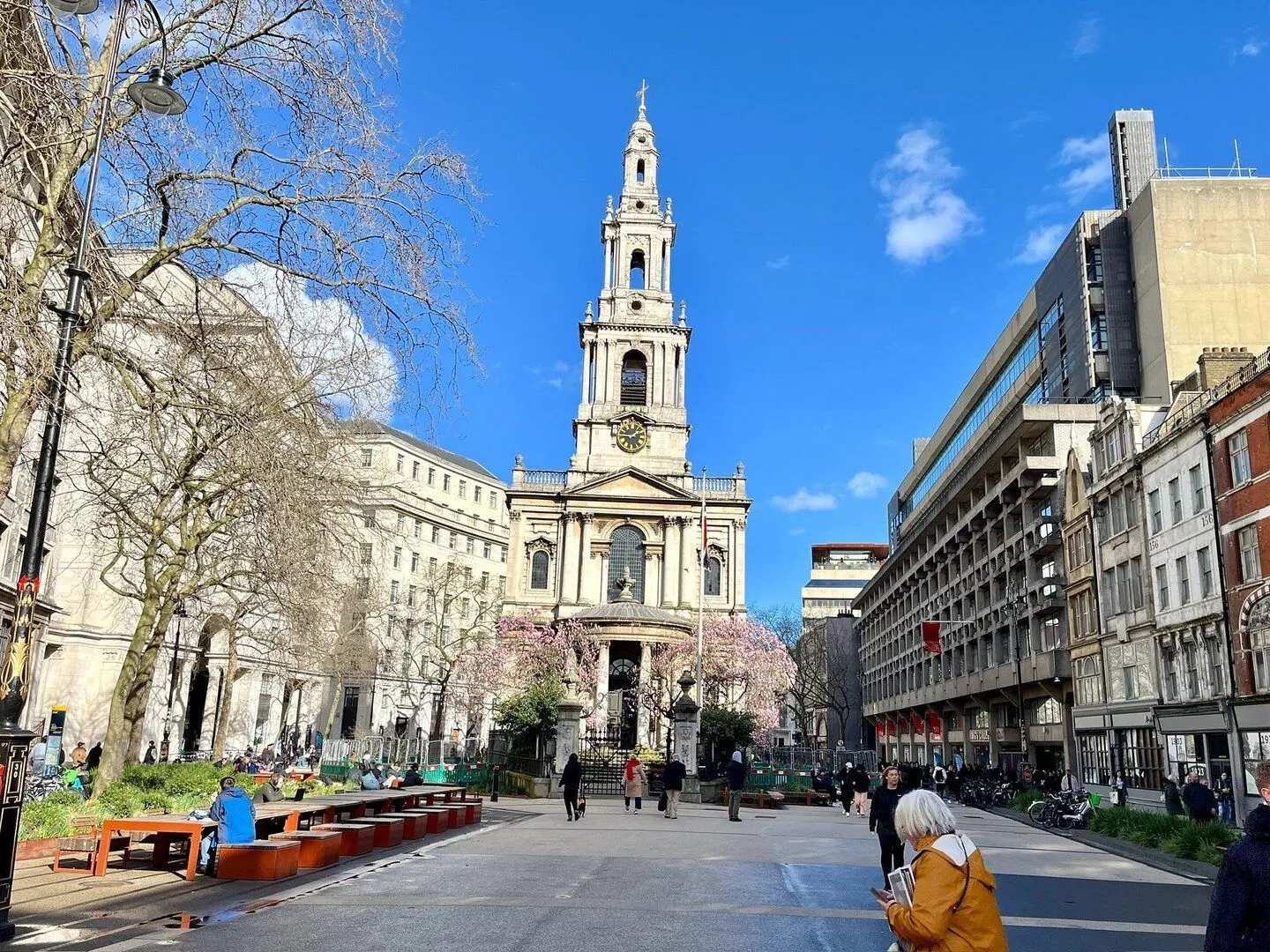An editorial image showcasing a historic church with a tall steeple, set within a bustling city square under a clear blue sky. People are visible walking and gathering in the foreground and midground, with blossoming trees adding a touch of color to the urban landscape.