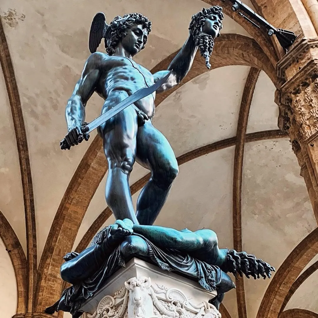A high-quality photograph of a bronze statue, likely Perseus with the Head of Medusa. The statue is well-composed within the frame, showing clear details and the architectural background provides good context without being distracting.