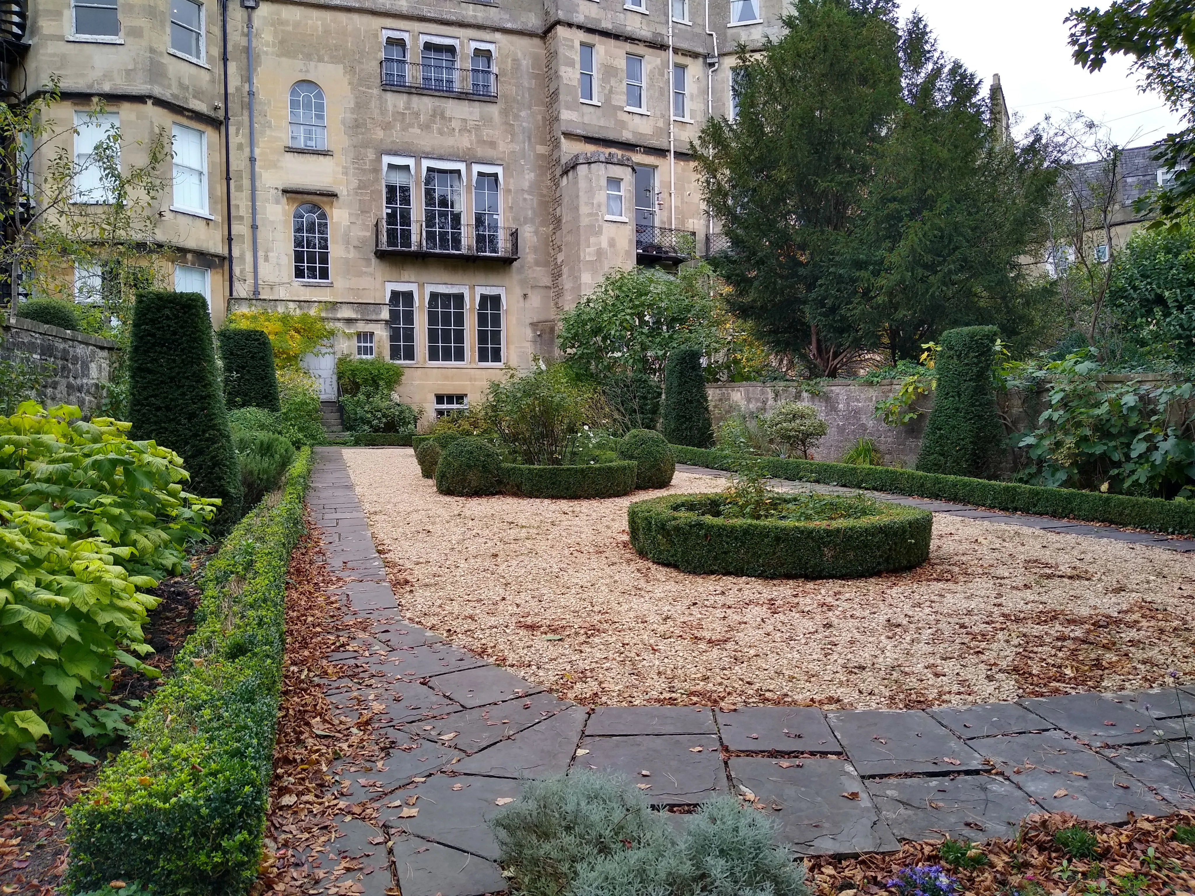 An editorial style image showcasing a well-maintained formal garden with structured hedges and gravel paths, set against a classic stone building under soft, natural lighting.