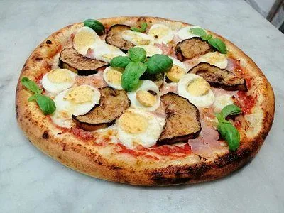 An overhead shot of a round pizza topped with sliced hard-boiled eggs, roasted eggplant, and fresh basil leaves, resting on a light-colored surface.
