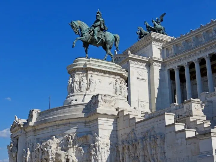 The image features a grand equestrian statue atop a large, ornate stone monument against a clear blue sky. The composition is strong, placing the statue prominently while also showcasing the intricate architectural details of the building. The lighting is excellent, illuminating the subject evenly and highlighting its textures and forms. The image is sharp, and colors appear natural and vibrant, especially the rich blue of the sky and the greenish patina of the bronze statue against the white stone. All key elements are in sharp focus, contributing to a high level of photo-realism.