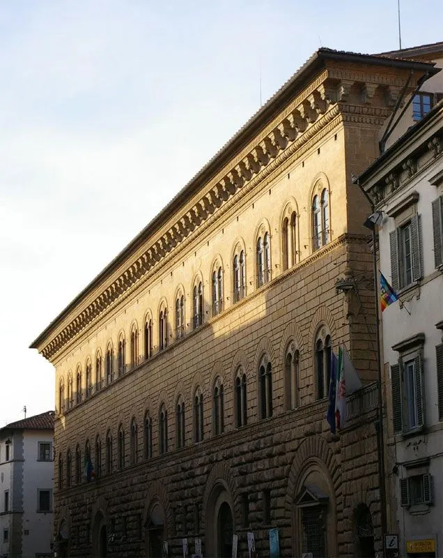 An editorial-style image depicting a large, historic stone building facade under sunlight. The building features multiple arched windows and intricate detailing. Another building is partially visible on the right, with flags hanging.