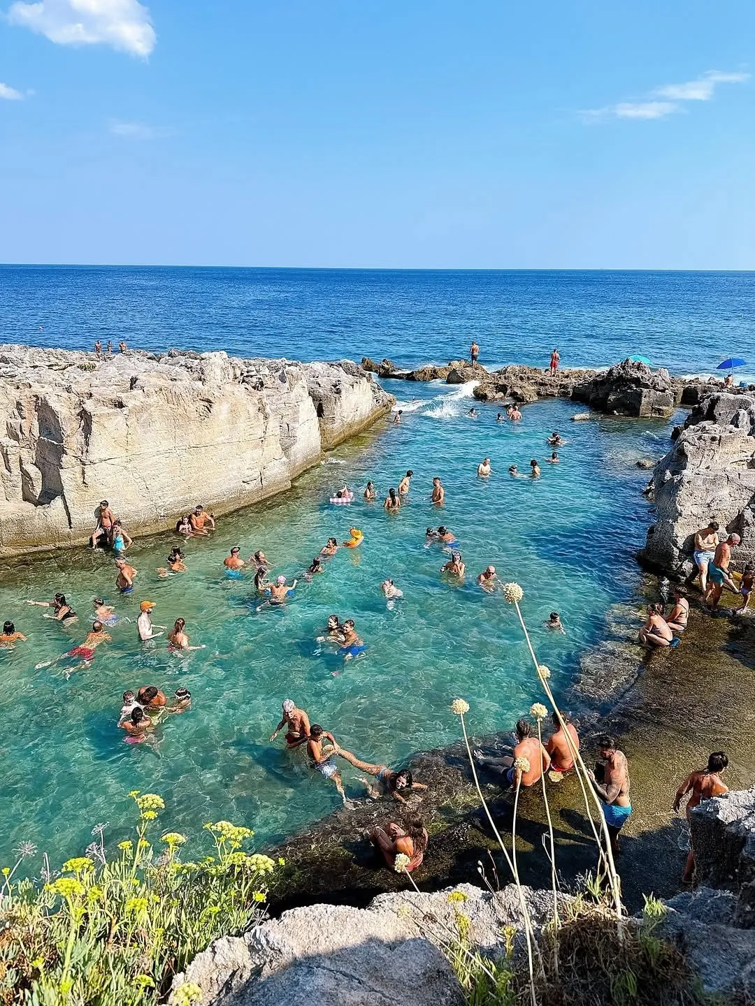A vibrant and realistic photograph of a natural rock pool filled with people swimming on a sunny day. The clear blue and turquoise water contrasts beautifully with the rocky formations.