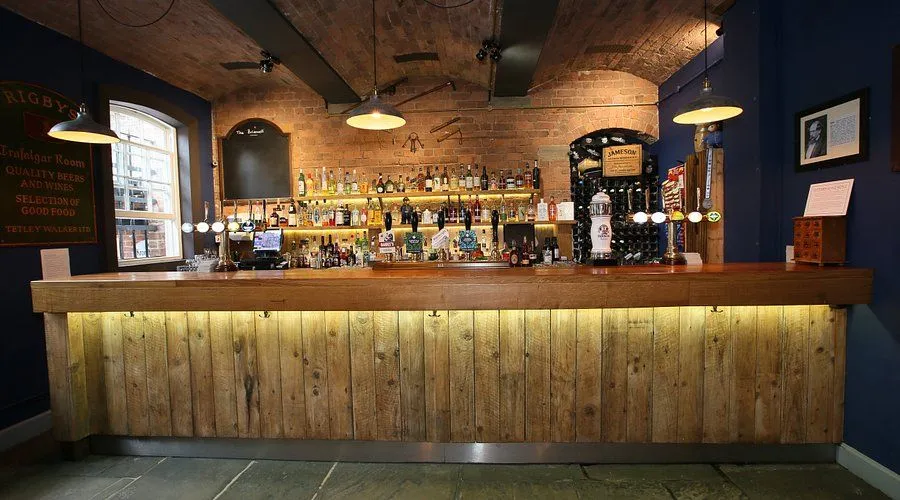 The image is well-composed, providing a clear, centered view of the bar. The lighting is generally good, highlighting the bar and shelves with a warm ambiance, though some upper areas are darker. The image is sharp, photo-realistic, and exhibits accurate colors, with all key elements, such as bottles and taps, being in clear focus.
