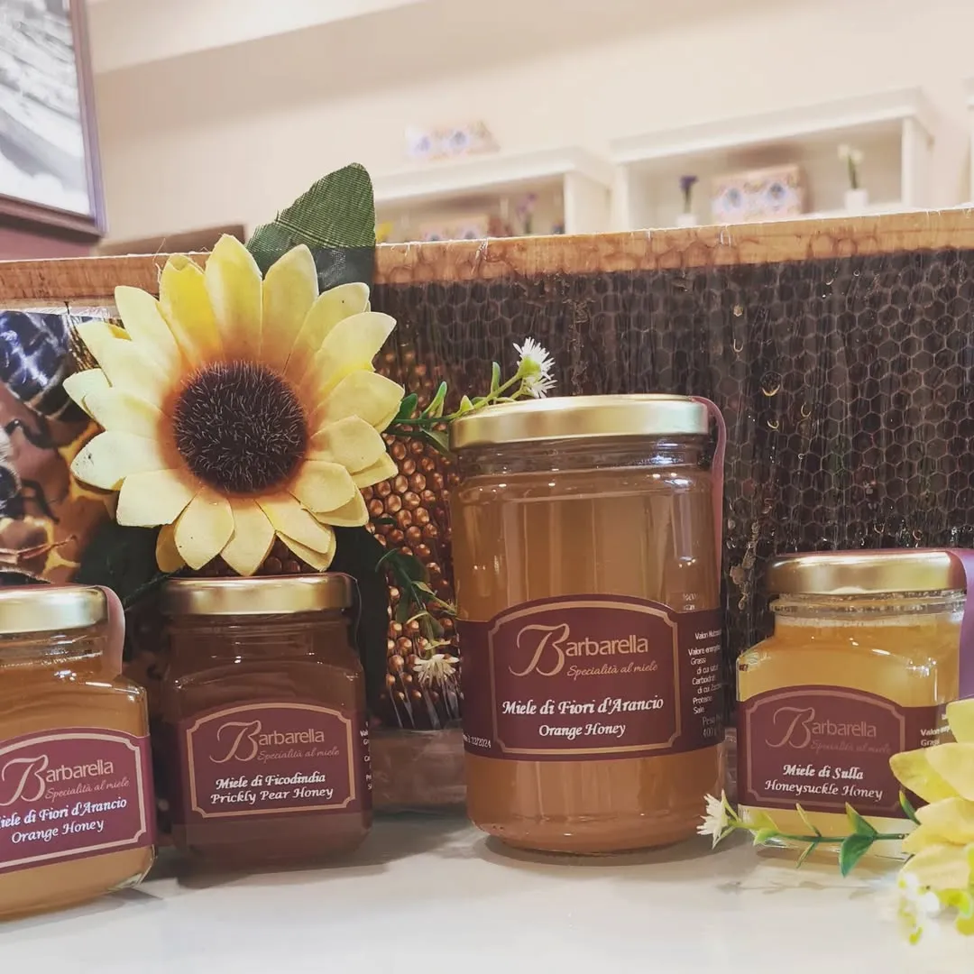 A well-composed and well-lit image featuring several jars of "Barbarella" honey, including orange, prickly pear, and honeysuckle varieties. The jars are arranged in the foreground with a sunflower and honeycomb in the background, creating a natural and appealing product display.