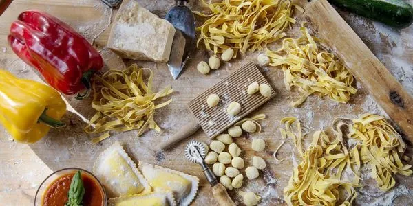 An overhead shot of various types of fresh pasta, including tagliatelle, ravioli, and gnocchi, alongside a block of Parmesan cheese and traditional pasta-making tools on a rustic wooden surface dusted with flour.
