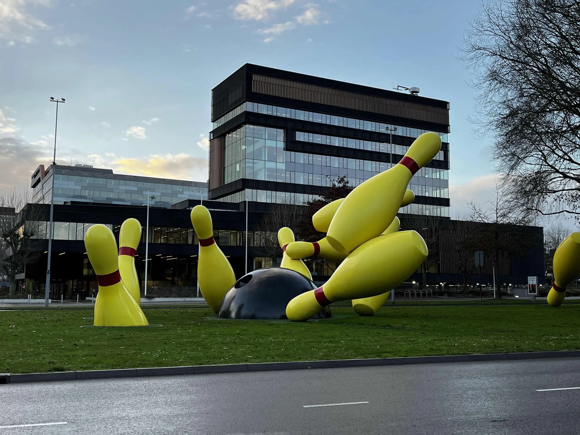 This image features a large outdoor sculpture depicting a bowling ball striking several bowling pins, set on a grassy patch with a modern building in the background under a cloudy sky. The colors are vibrant and true to life.