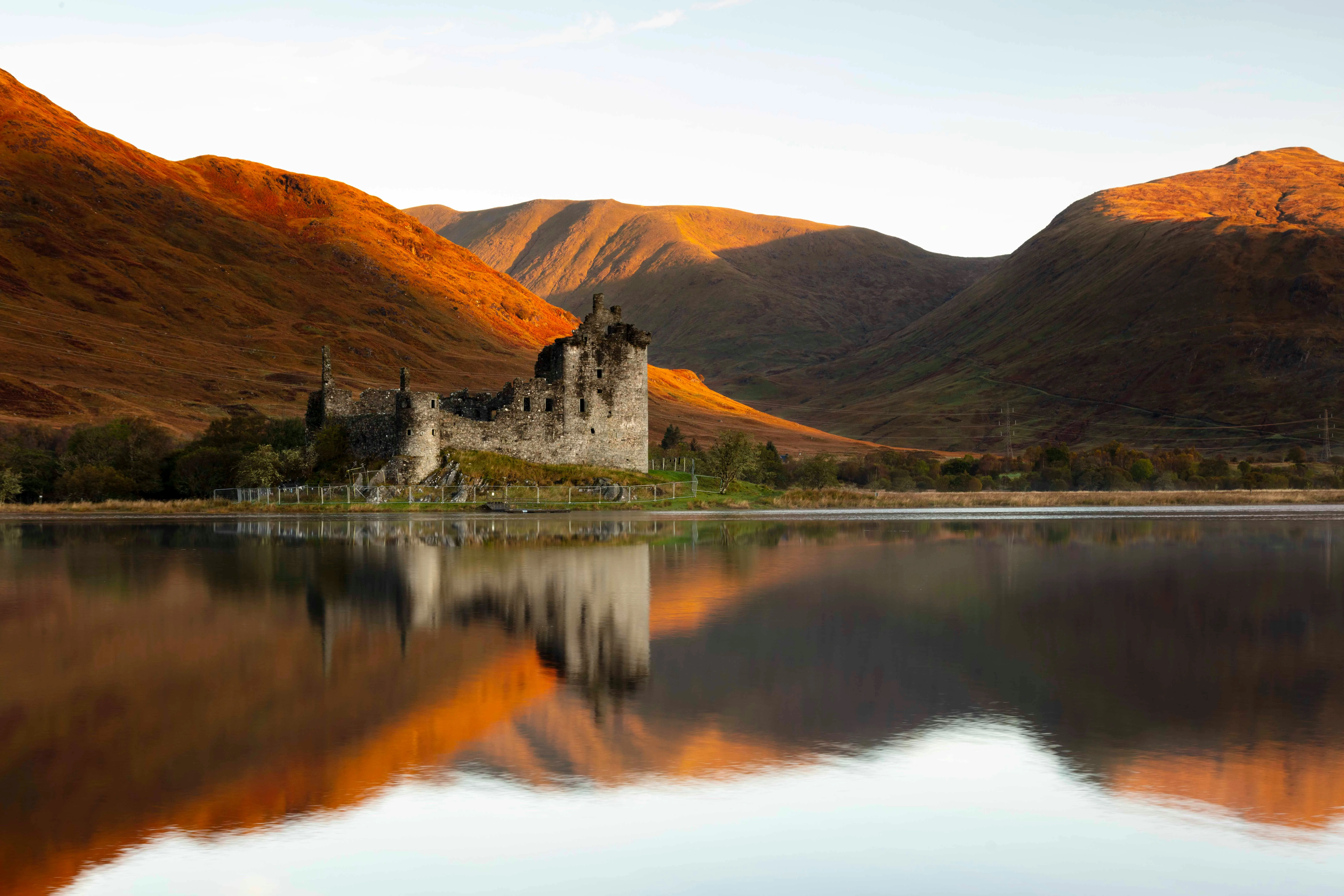 A stunning landscape photograph featuring the ruins of Kilchurn Castle reflecting in a tranquil loch, set against a backdrop of mountains illuminated by the warm light of sunrise or sunset.