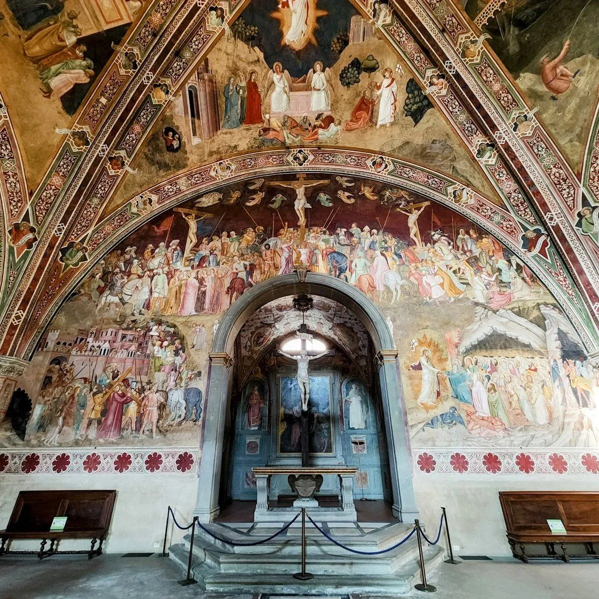An interior shot of a chapel adorned with detailed religious frescoes covering the walls and ceiling, focusing on an altar at the center. The frescoes depict various biblical scenes with multiple figures.