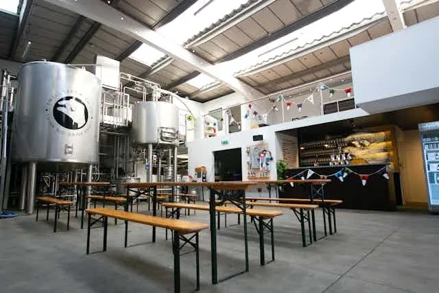 The image captures a well-composed interior view of a brewery or taproom, featuring stainless steel tanks and casual wooden tables with benches. The scene is brightly lit, creating a clear and photo-realistic representation. Key elements in the foreground and mid-ground are sharp and in focus, and the colors are accurate. While generally well-lit, some areas of the ceiling appear slightly overexposed.