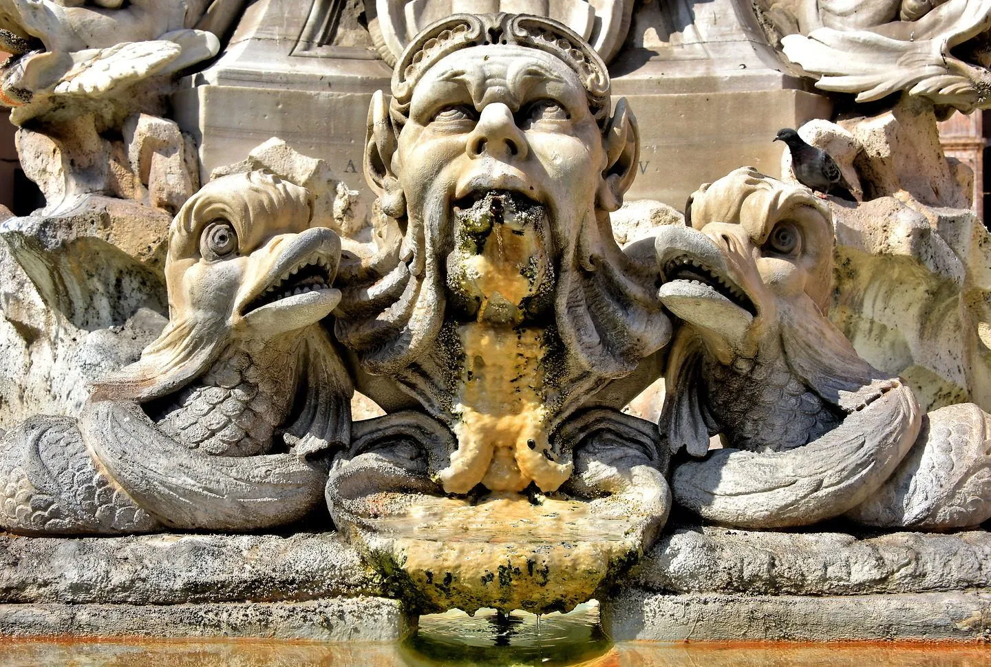 A detailed close-up of an ornate stone fountain featuring a central gargoyle-like figure spouting water, flanked by two other mythical creatures. The water flow and the weathered texture of the stone are prominent.