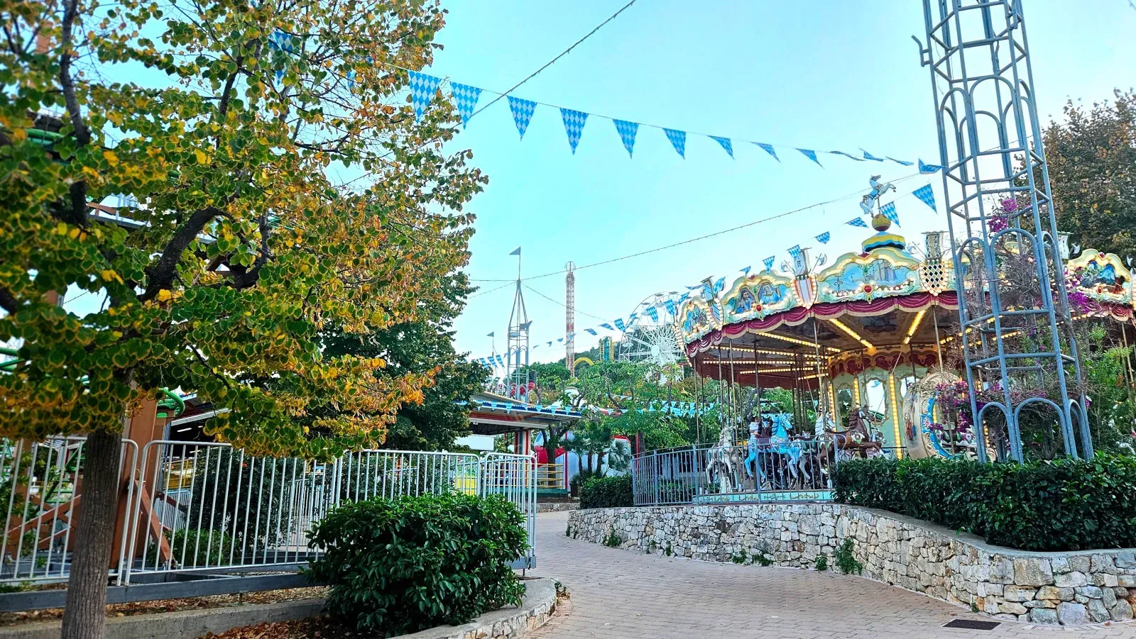 An outdoor scene at an amusement park featuring a colorful carousel, festive checkered pennants strung across the sky, and various other rides in the background, including a Ferris wheel. A large tree frames the left side of the image, and a paved path leads into the scene.