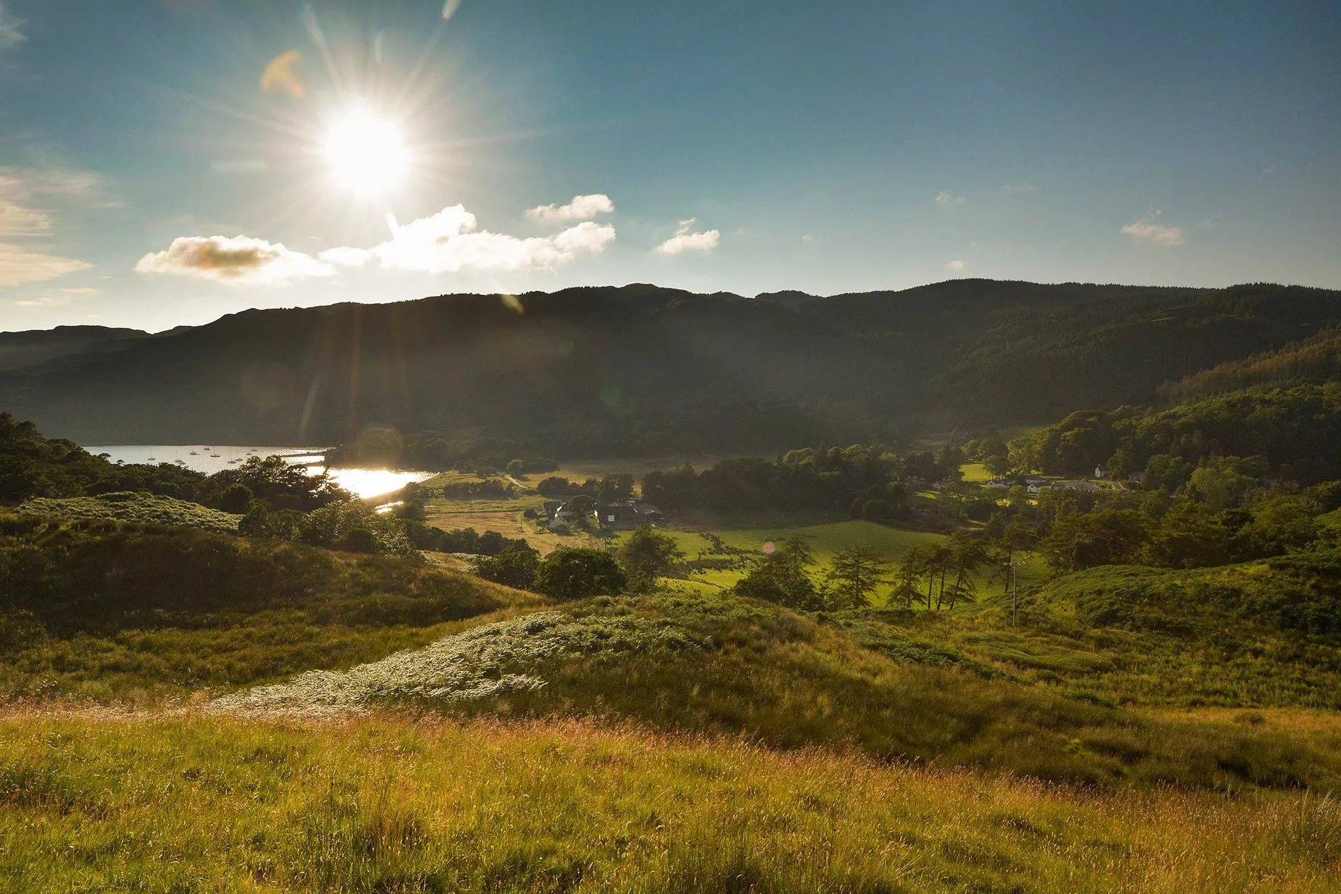 A scenic landscape featuring rolling hills with golden grass in the foreground, leading into a valley with a body of water reflecting the sun. Distant mountains frame the background under a blue sky with scattered clouds, with a strong sunburst emanating from the upper left.