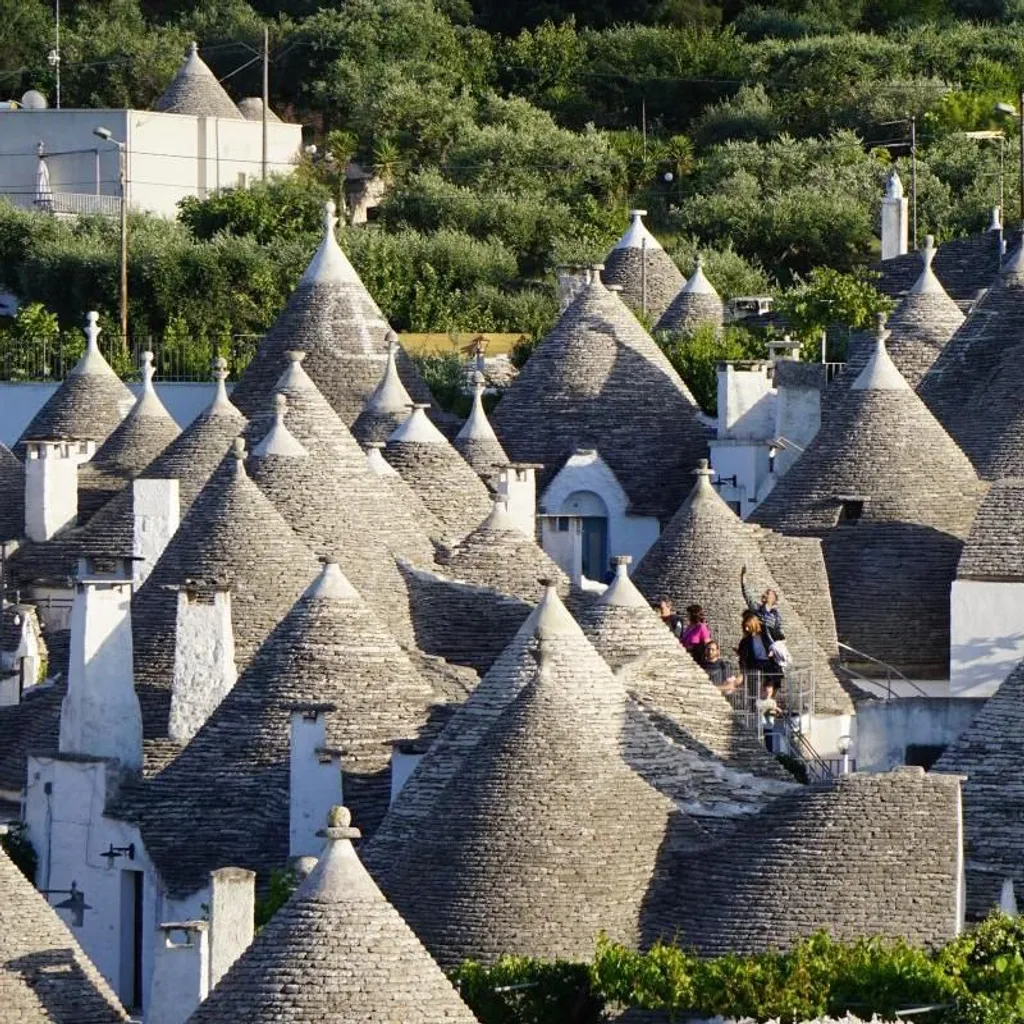 An elevated view showcasing a dense cluster of traditional trulli houses with their distinctive conical stone roofs, set amidst green foliage in Puglia, Italy.