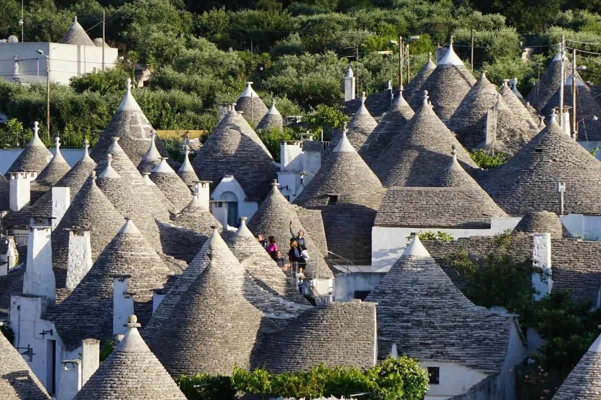 An elevated view showcasing a dense cluster of traditional trulli houses with their distinctive conical stone roofs, set amidst green foliage in Puglia, Italy.